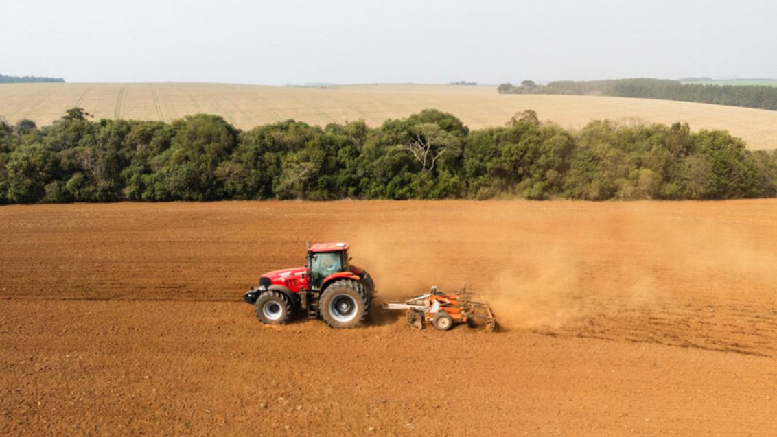 Rode tractor op akker met stofwolk, bomenrij op de achtergrond